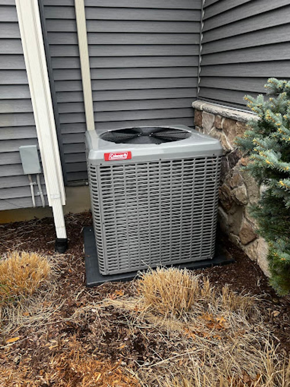 Gray outdoor air conditioning unit near a stone wall and dry landscaping.