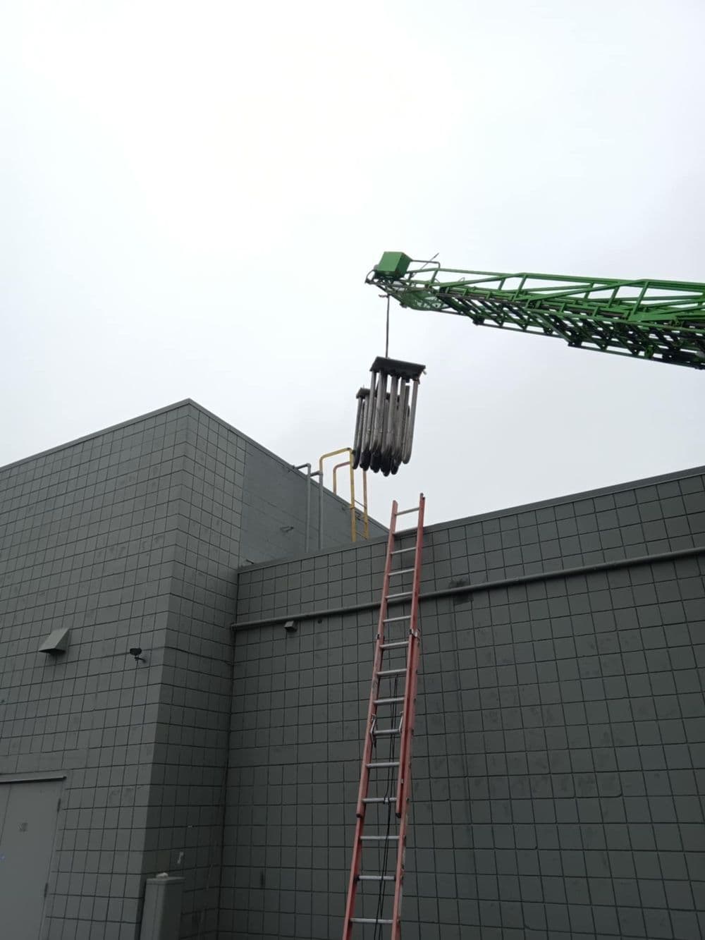 Green crane lifting pipes to rooftop with ladder against gray building.