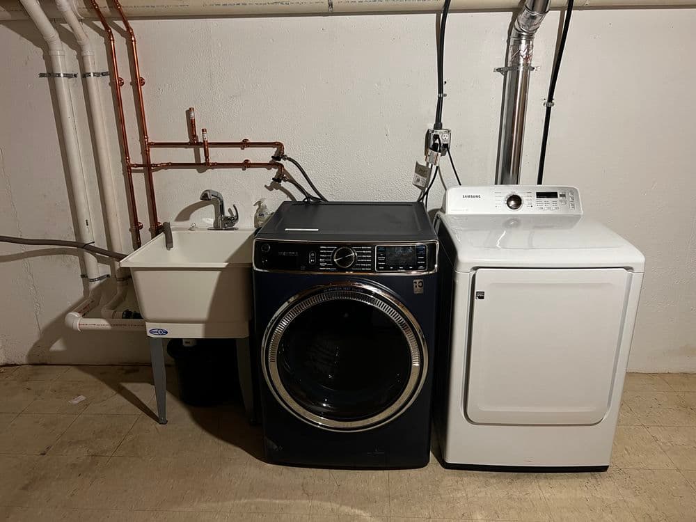 Washer and dryer set in a basement laundry area next to a utility sink with plumbing.