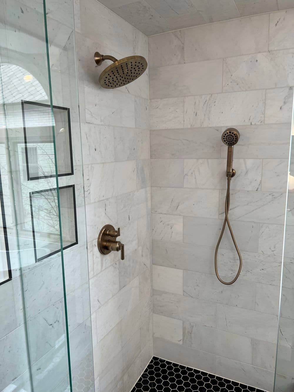 Modern shower with dual bronze fixtures, marble walls, and black hexagonal tile floor.
