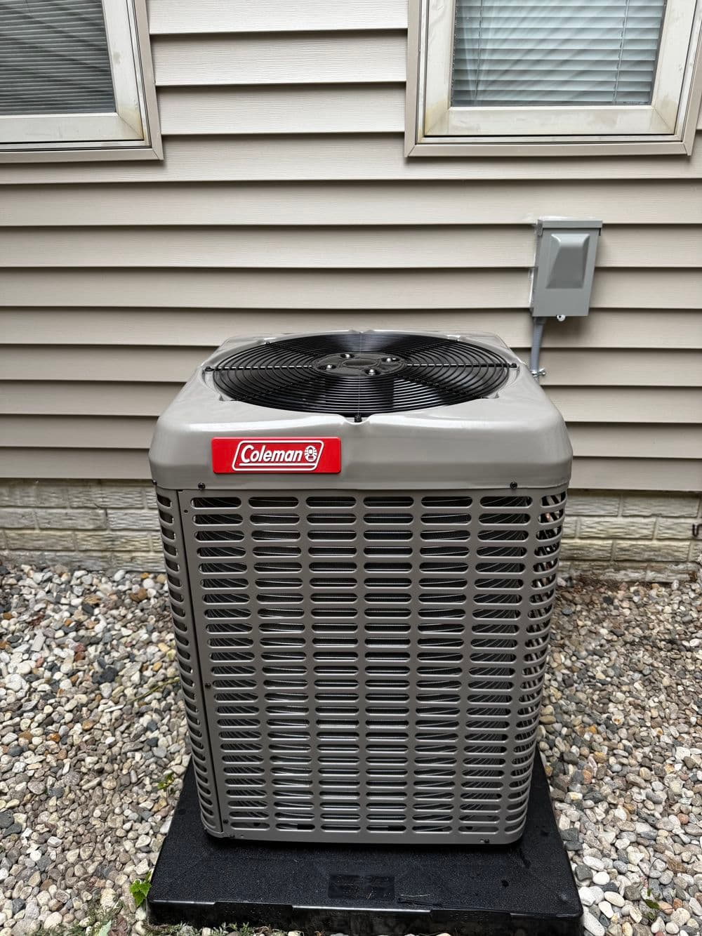 Coleman air conditioning unit outside a home with beige siding and gravel ground.
