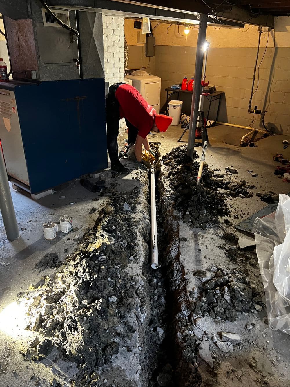 Worker repairing plumbing in a basement with exposed pipes and a trench dug in the floor.